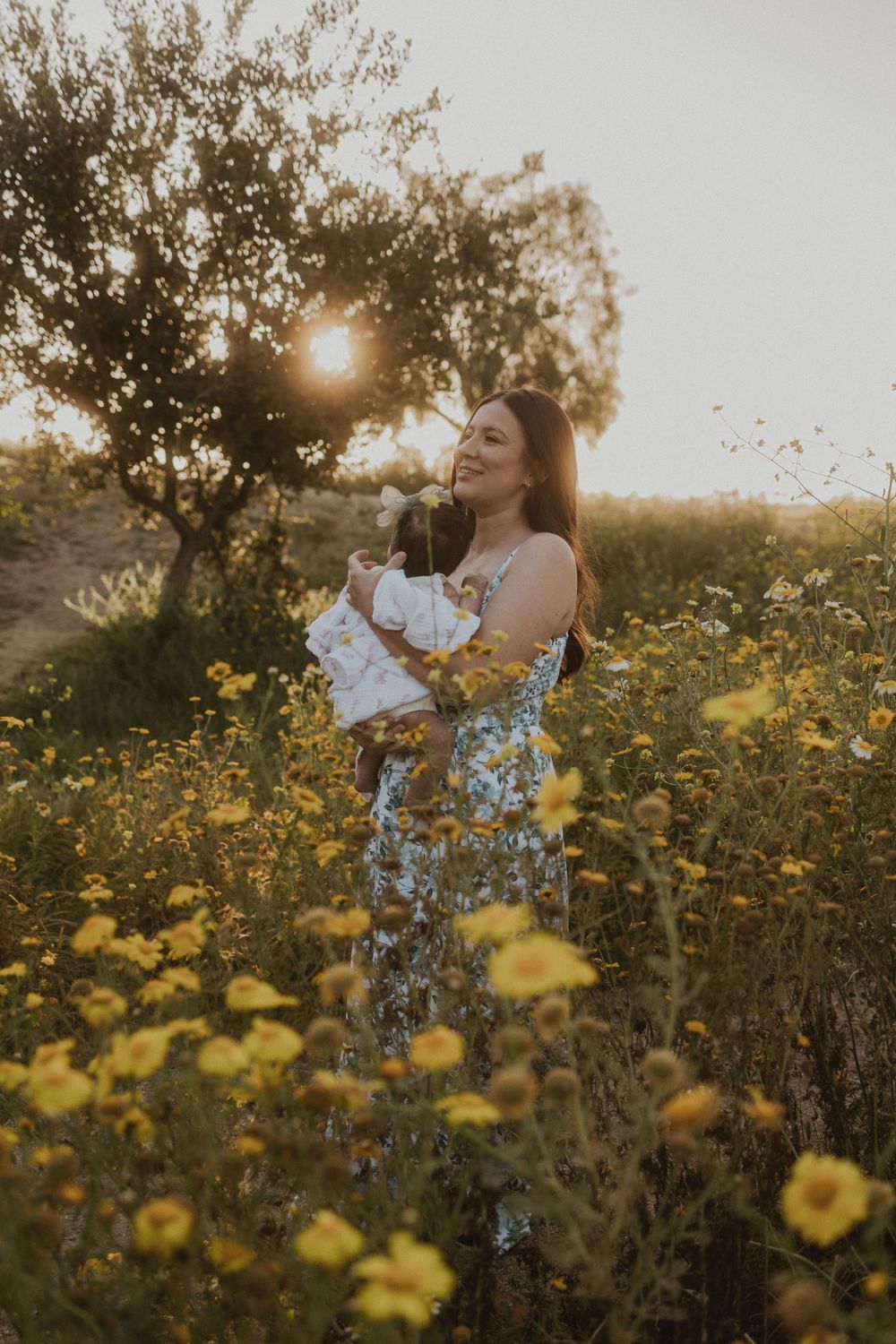 Woman in a floral dress holding a baby, standing in a field of yellow flowers at sunset.