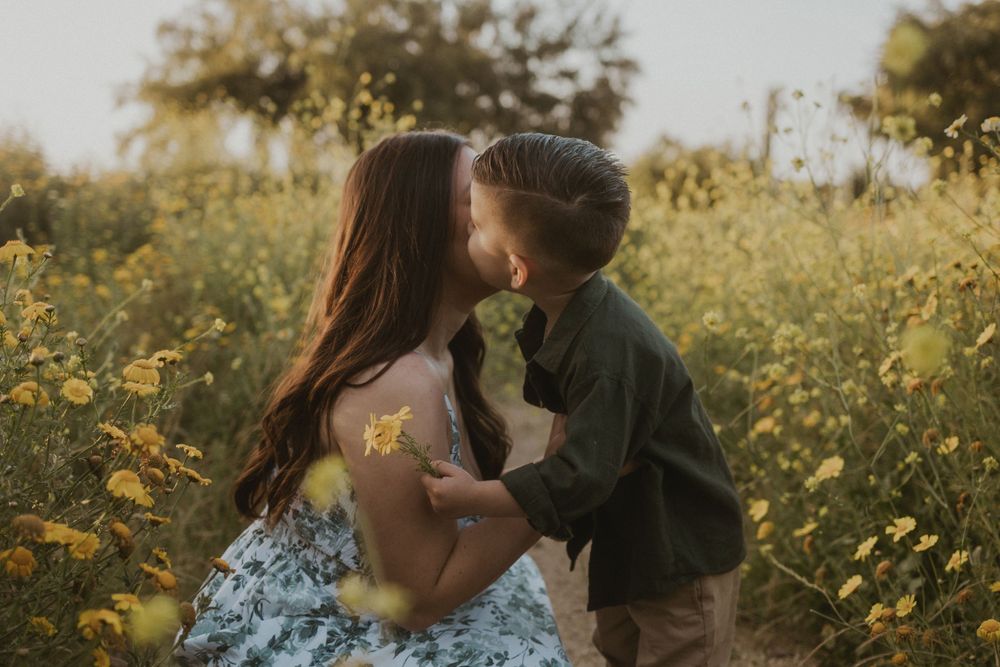 A woman and a young boy kissing in a field of yellow wildflowers. The woman holds a small bouquet of yellow flowers.