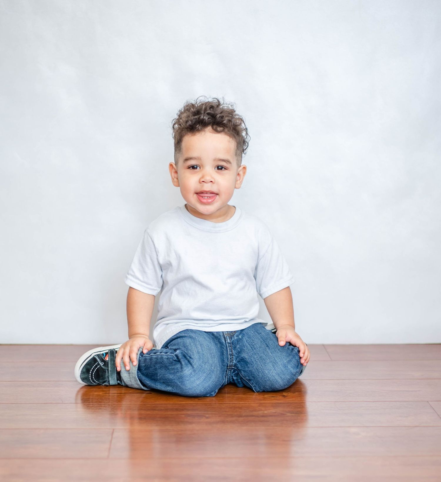 A toddler in white shirt and jeans sits casually on hardwood flooring against a plain background.