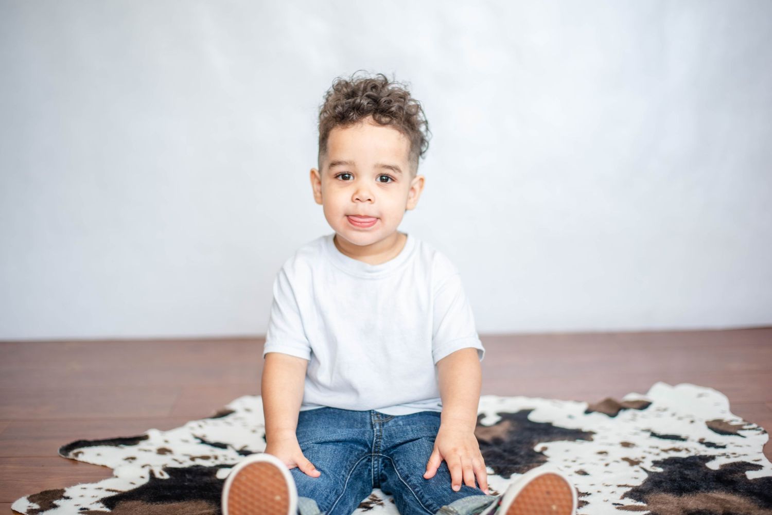 Child sits on wooden floor next to black and white cowhide rug in bright studio setting.