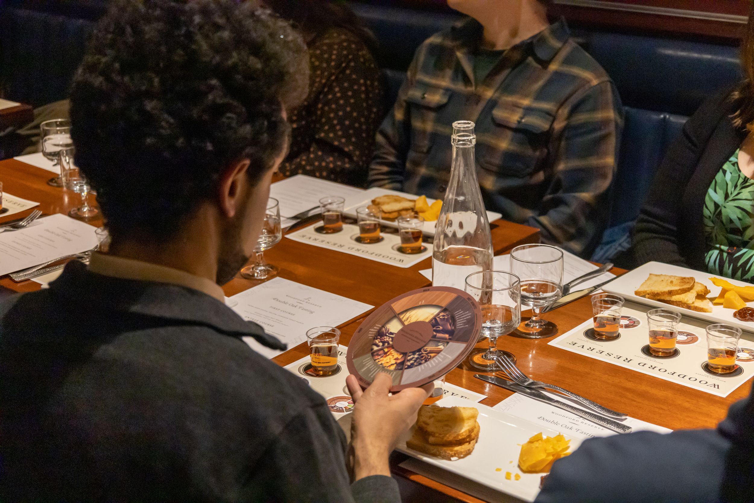 People gathered around a wooden table for a tasting event with food and drinks.