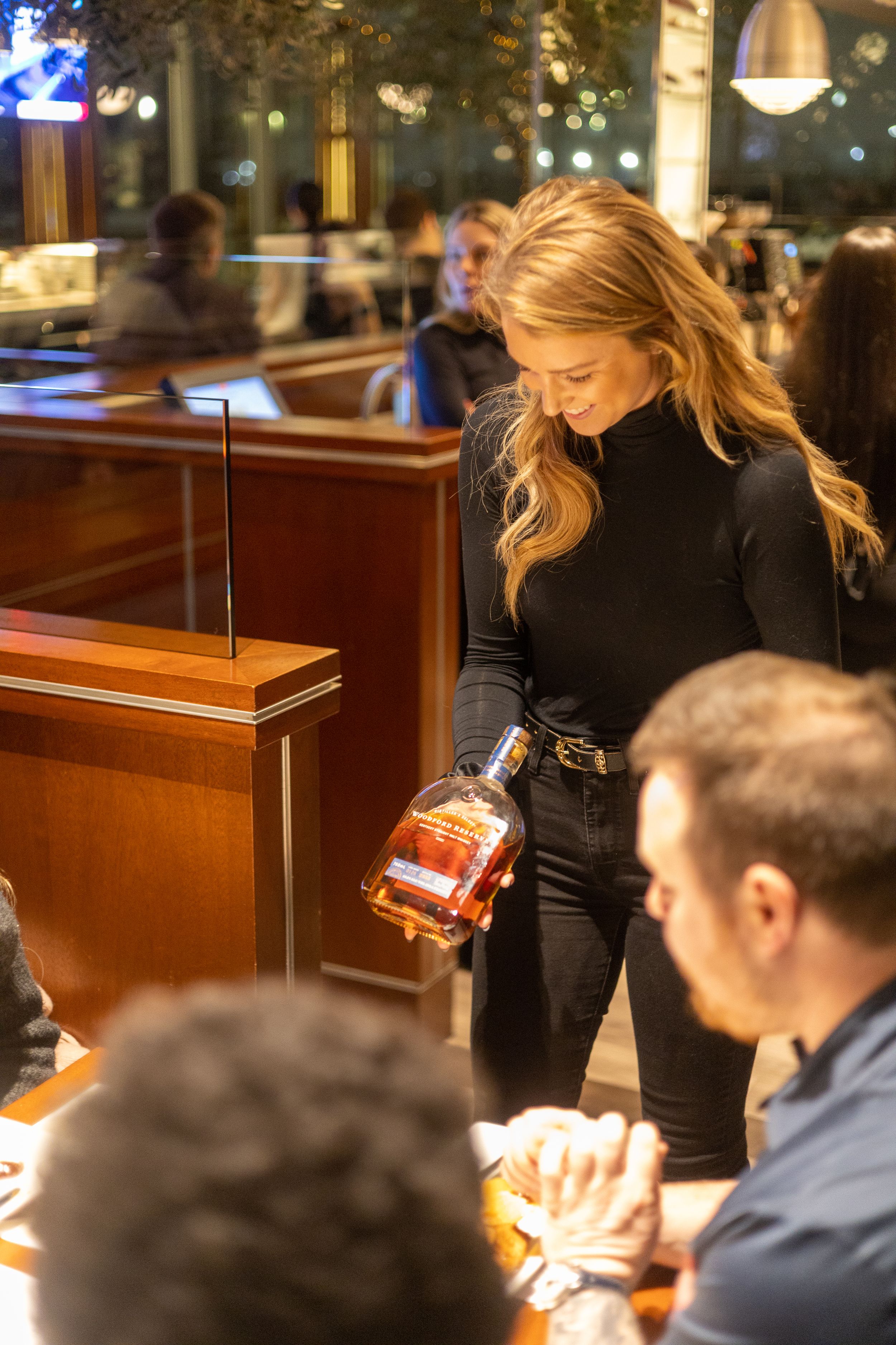 Server in black attire presenting a bottle of spirits to diners at an upscale restaurant.