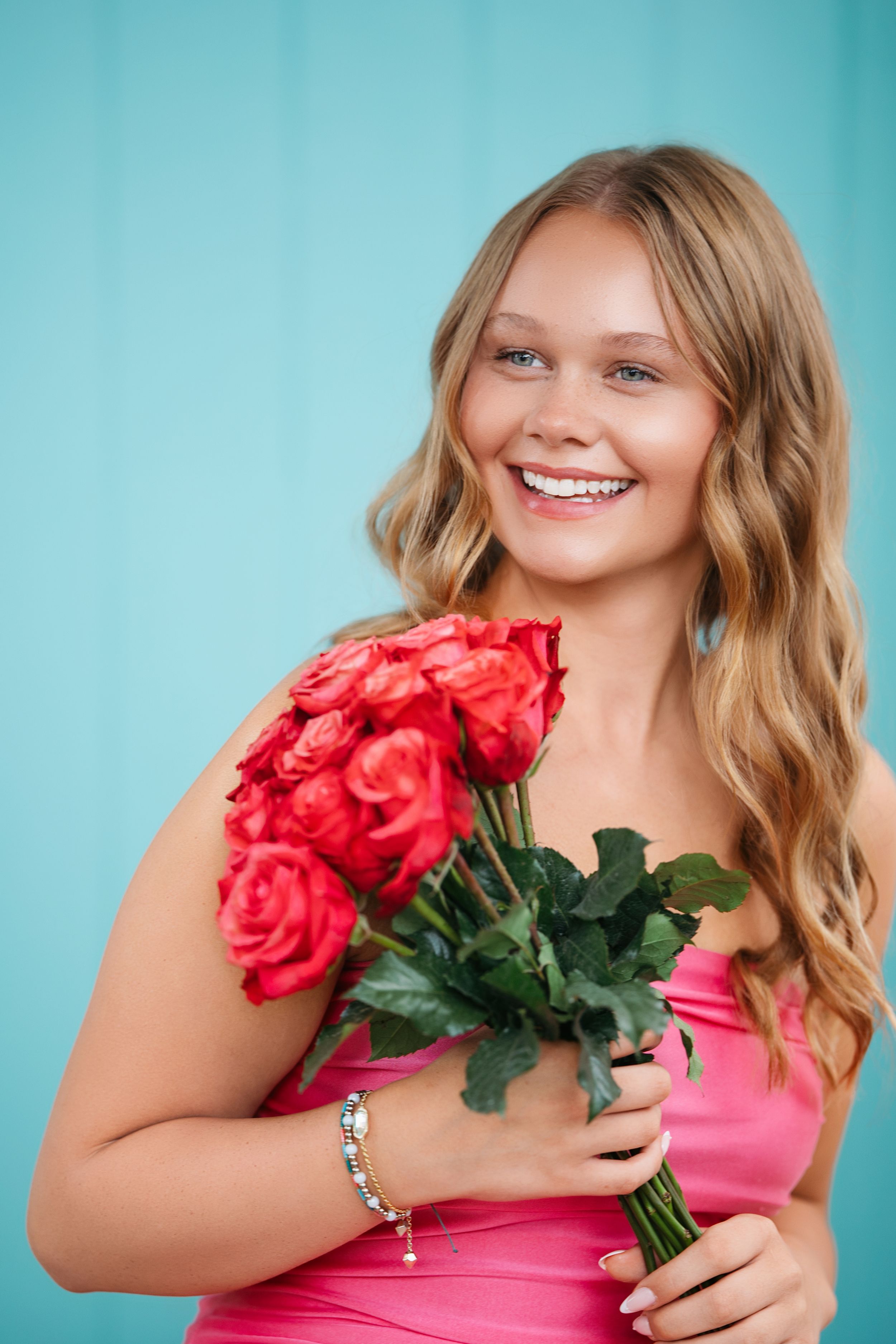 Senior girl middle Tennessee in pink dress poses with red roses against turquoise background in fun portrait series.