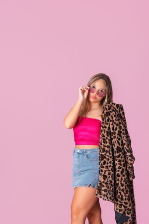 Woman in pink crop top and denim shorts poses playfully with leopard print coat against pink background.