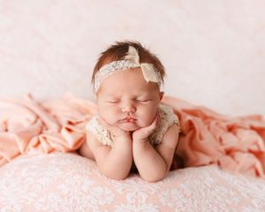 A sleeping newborn baby wearing a delicate lace headband lies peacefully on soft pink fabric.