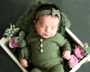 Newborn baby sleeping peacefully in green knit outfit with pink roses on white wooden prop bed.