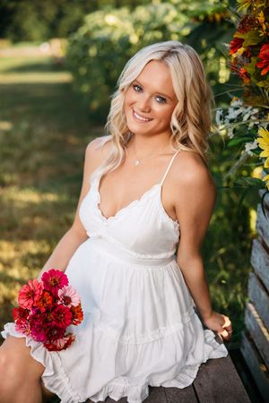 A woman in a white summer dress poses outdoors with red carnations against a natural garden backdrop.