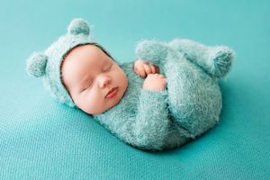 A sleeping baby wearing a teal knit hat lies on a teal background with matching plush toy.