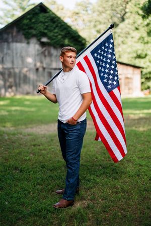 A patriotic scene with someone holding an American flag in front of a rustic barn on a sunny day.