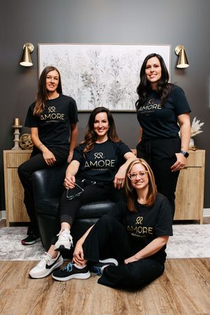 Group wearing matching black Amore branded t-shirts and pants sit casually on wooden floor against floral wallpaper.