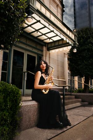 A musician in a black dress sits on outdoor steps playing a saxophone in a moody architectural setting.
