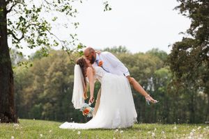 Bride in white dress twirling playfully in a sunny green meadow with trees in the background.