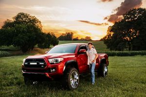 Red Toyota Tacoma pickup truck parked in grassy field during golden sunset with trees in background.