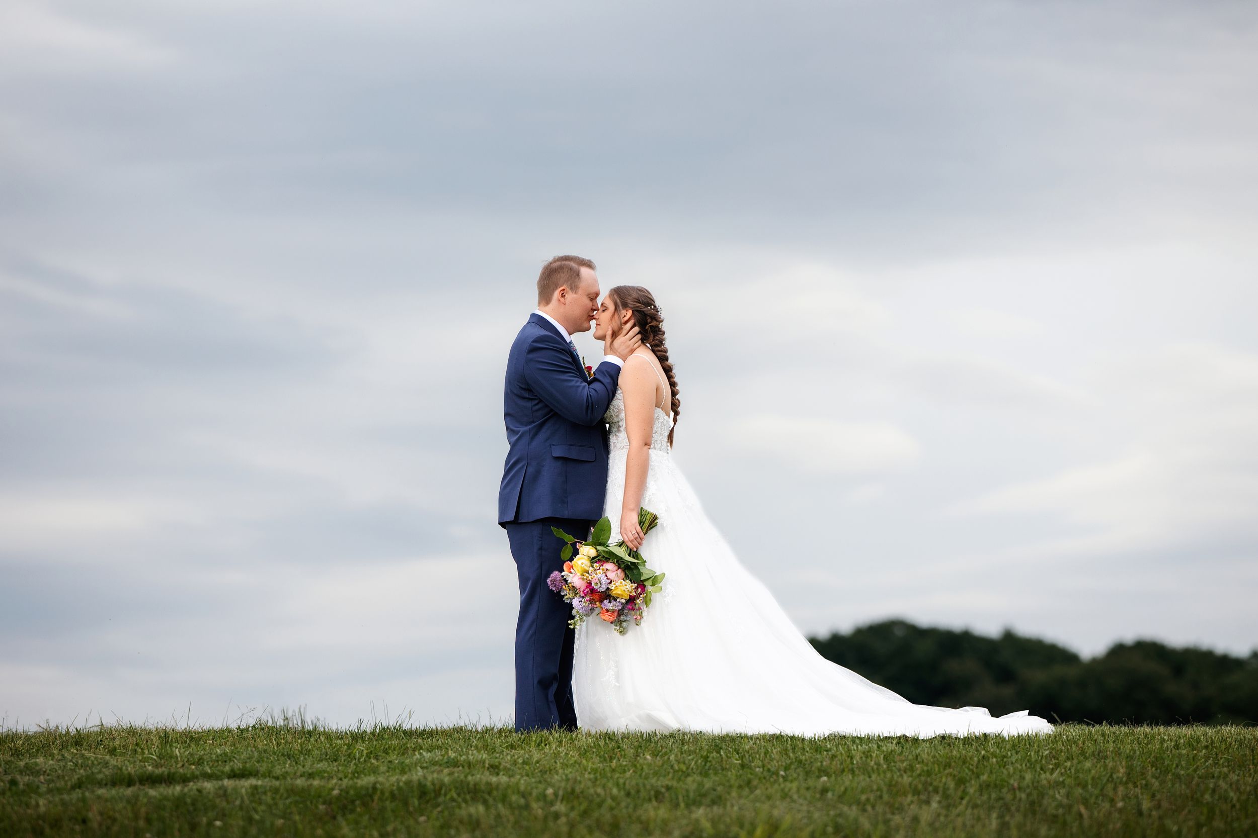 A couple in wedding attire shares a romantic moment on a hilltop against a cloudy sky.