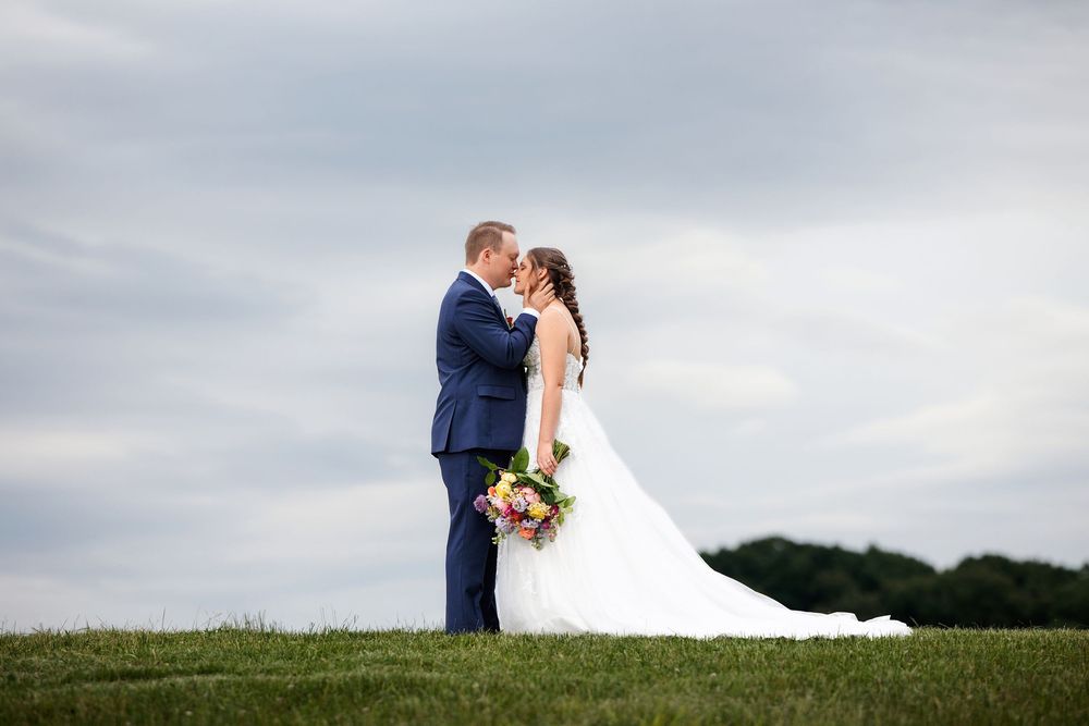 A couple in wedding attire shares a romantic moment on a hilltop against a cloudy sky.