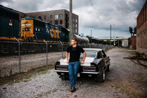 A person stands next to parked cars on a gravel road with industrial buildings in the background.