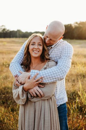 A couple embraces in a golden field at sunset, wearing casual attire with a blue checkered shirt and beige dress.
