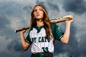 Softball player in green and white uniform holding bat over shoulder against stormy sky.