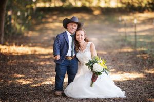 A romantic couple in wedding attire sits together on fallen autumn leaves in a sunlit forest setting.