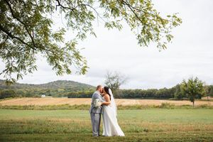 Wedding couple poses in a scenic field with trees and rolling hills in the background during golden hour.