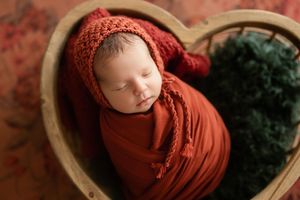 A sleeping newborn swaddled in rust-colored fabric lies peacefully in a wooden bowl with greenery accents.
