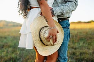 Romantic couple in western attire holds hands in a golden sunlit meadow during sunset.