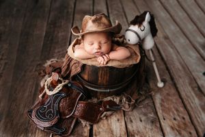 A sleeping newborn in a rustic cowboy themed setting with western boots and hat on wooden floorboards.