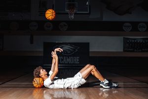 A basketball player lies on the court holding a ball while another ball hovers in mid-air above.