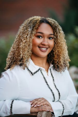 Professional portrait of someone with curly blonde hair wearing a white blouse with black trim and a warm smile.