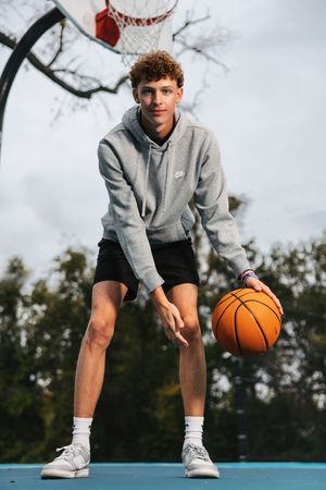 Basketball player in grey hoodie and shorts crouches on outdoor court with orange ball.