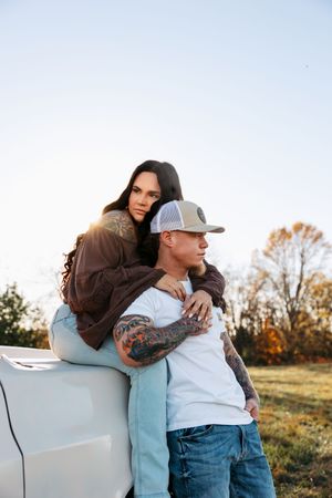A couple embraces while sitting on a car hood during sunset in an autumn field.