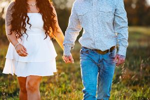 Couple holding hands while walking in a sunlit meadow, with white dress and blue jeans visible.