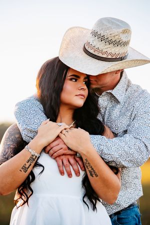Romantic western-style couple embracing outdoors at sunset, wearing cowboy hat and white dress with matching floral print shirt.