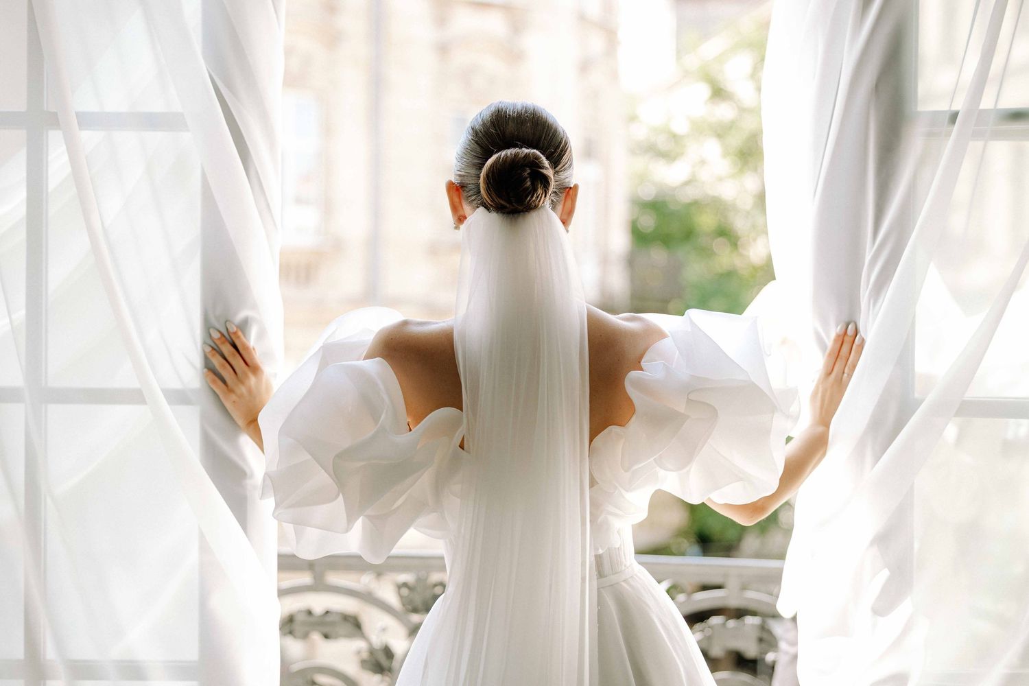 Elegant bride in white gown holds curtains while standing between railings outdoors.