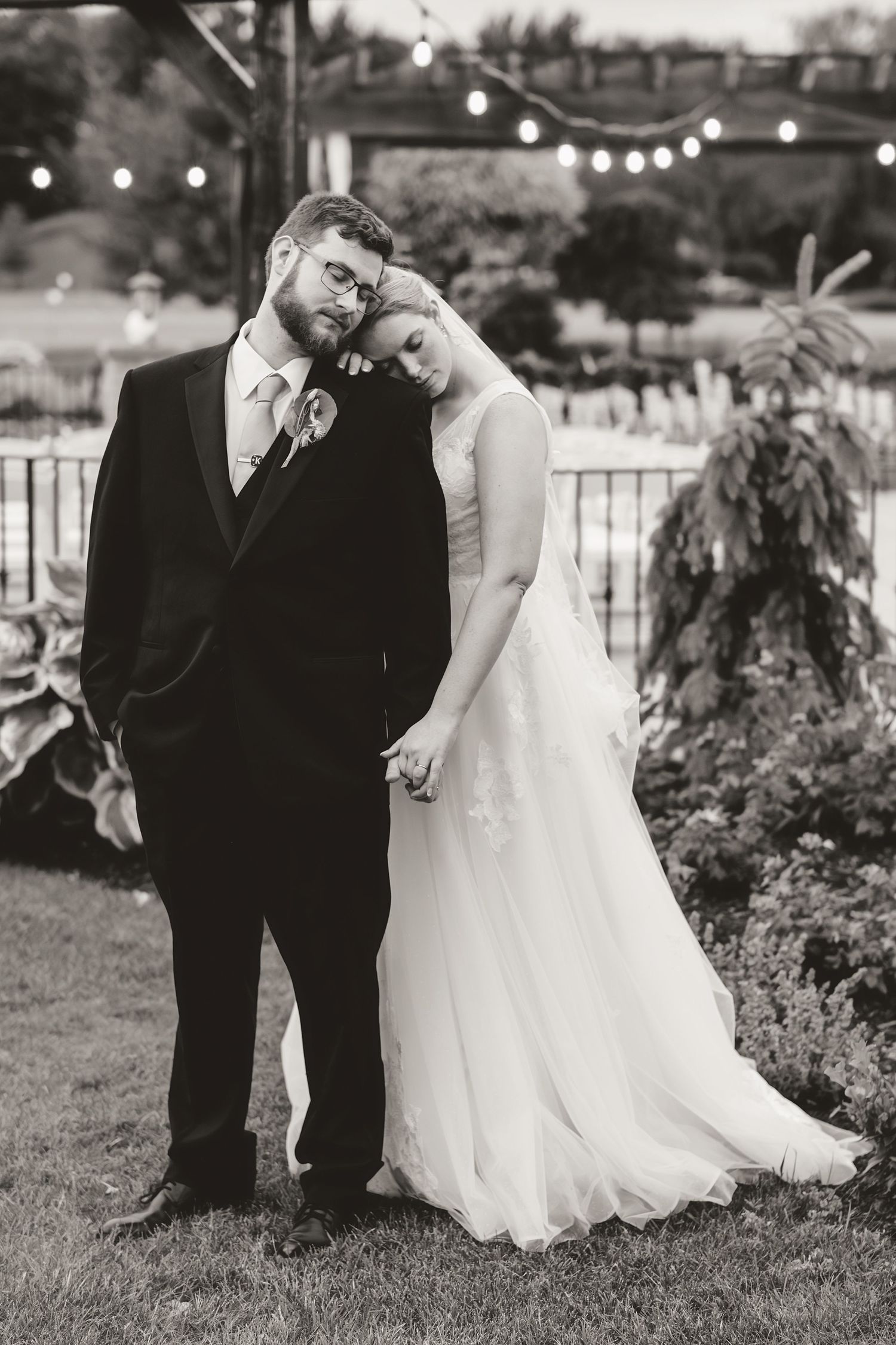 Black and white portrait of couple in wedding attire posing under string lights.