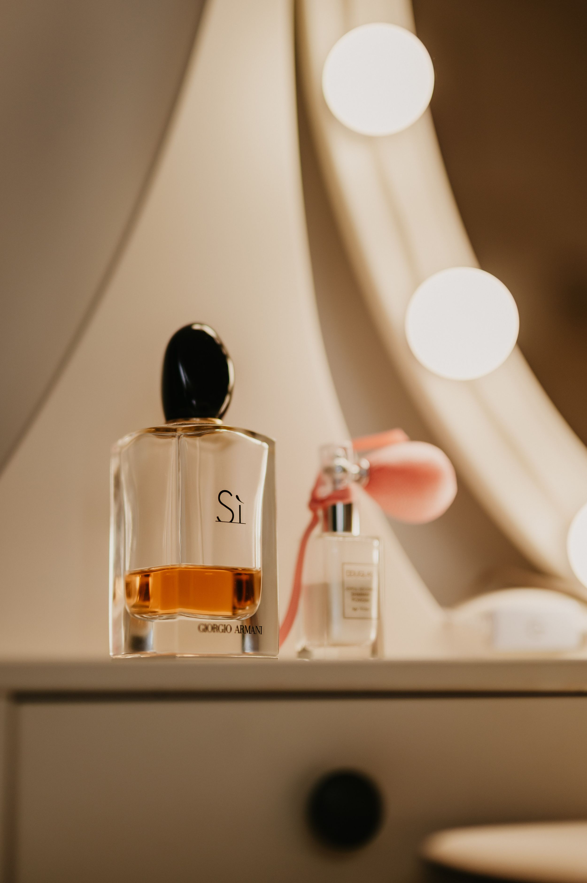 Perfume bottles with amber liquid sit on a vanity with soft bokeh lights in the background.