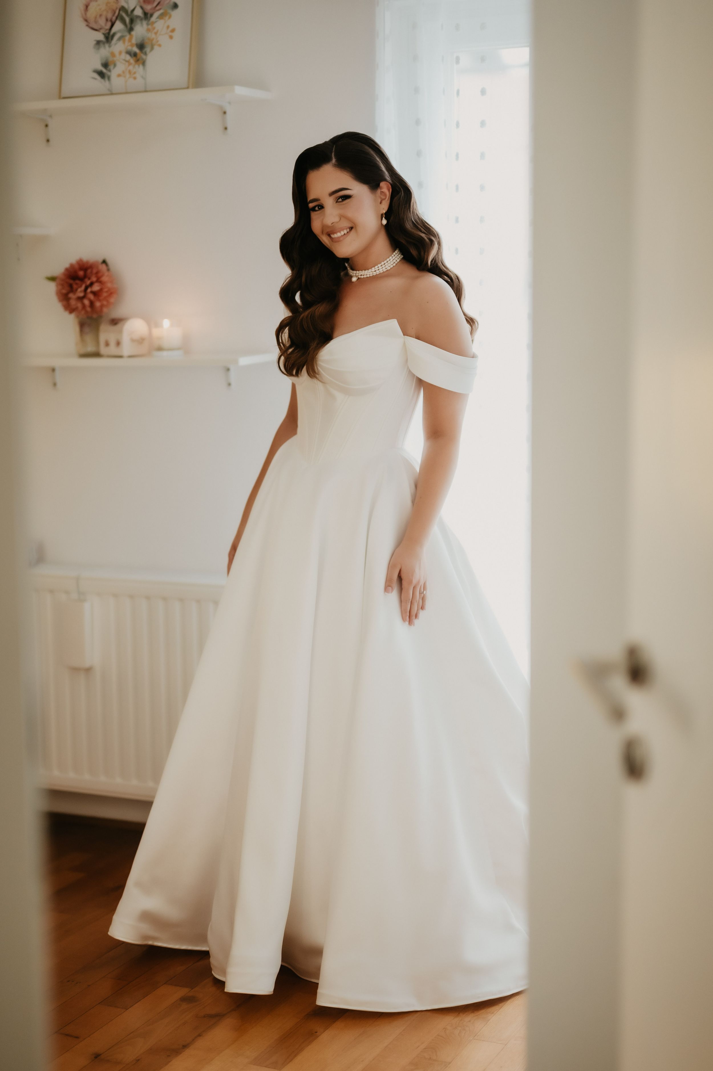 Elegant white wedding dress with off-shoulder design shown in a bright interior setting with soft decor details.