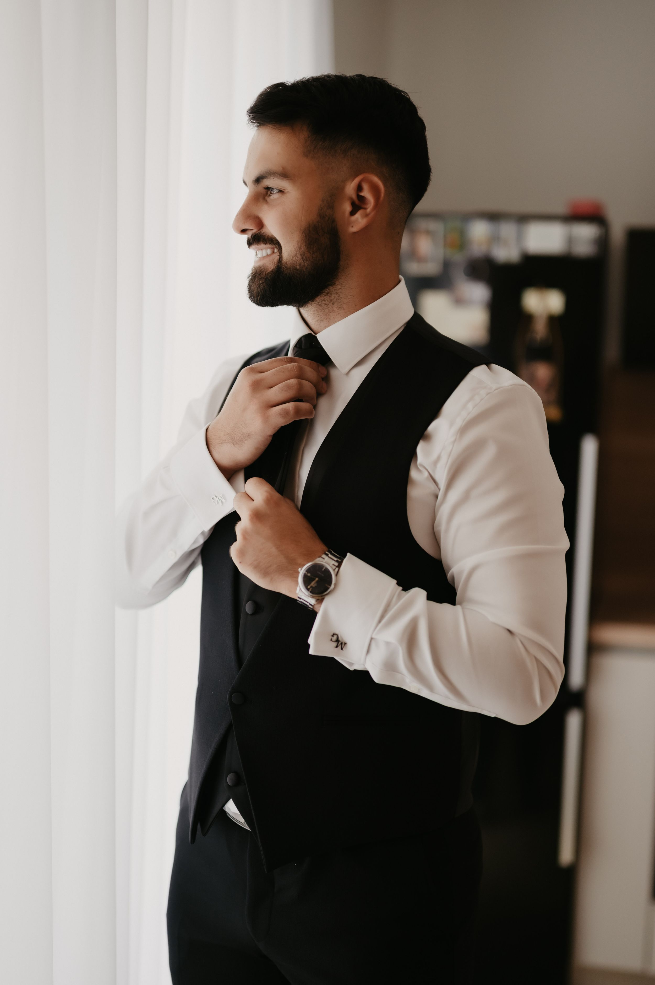 A groom in a black vest and white dress shirt thoughtfully touches his chin while getting ready for his wedding day.