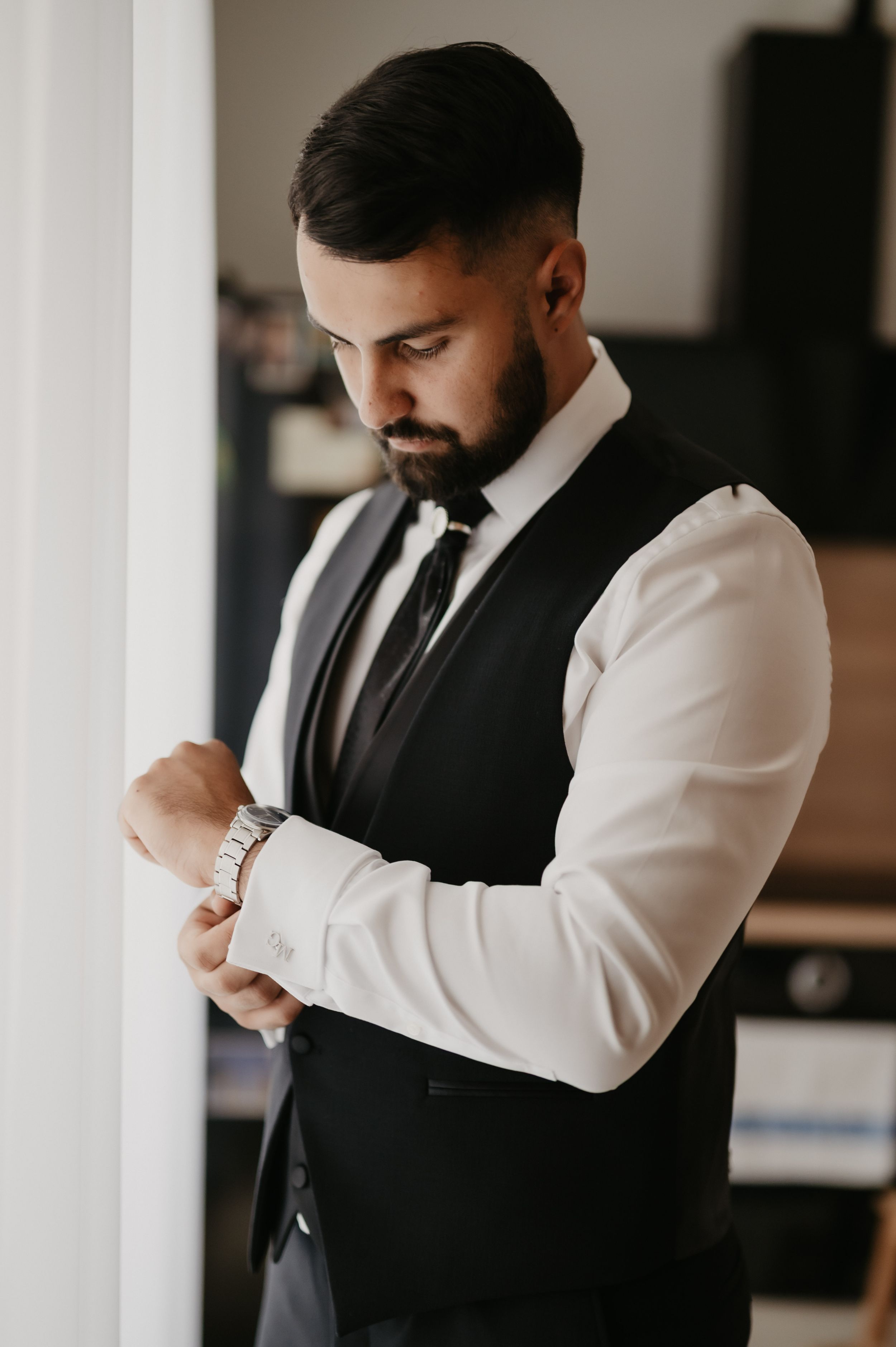 A groom in a black vest and white dress shirt adjusts his cuff links during wedding preparations.