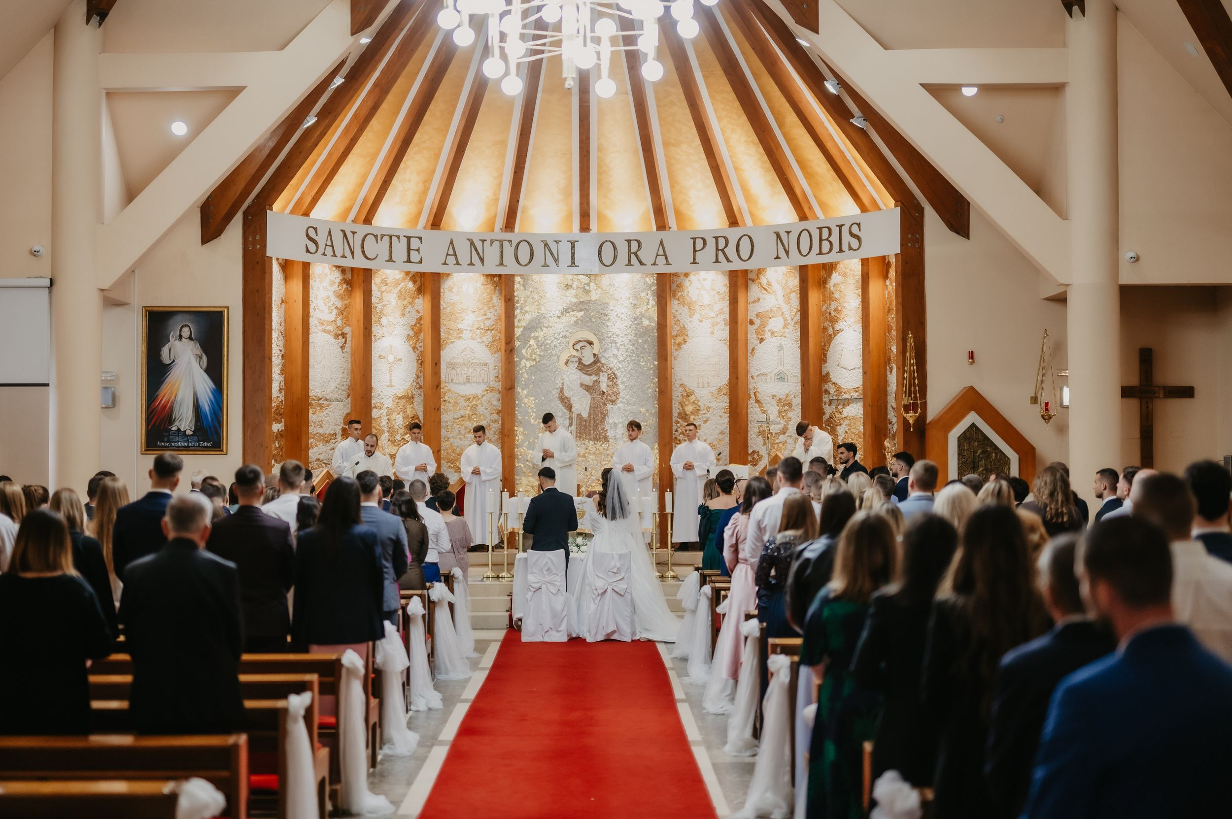 A traditional Catholic church wedding ceremony with a red carpet aisle and guests seated in wooden pews.