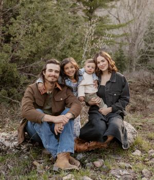 A family sits together on a rock in a mountain setting surrounded by trees.