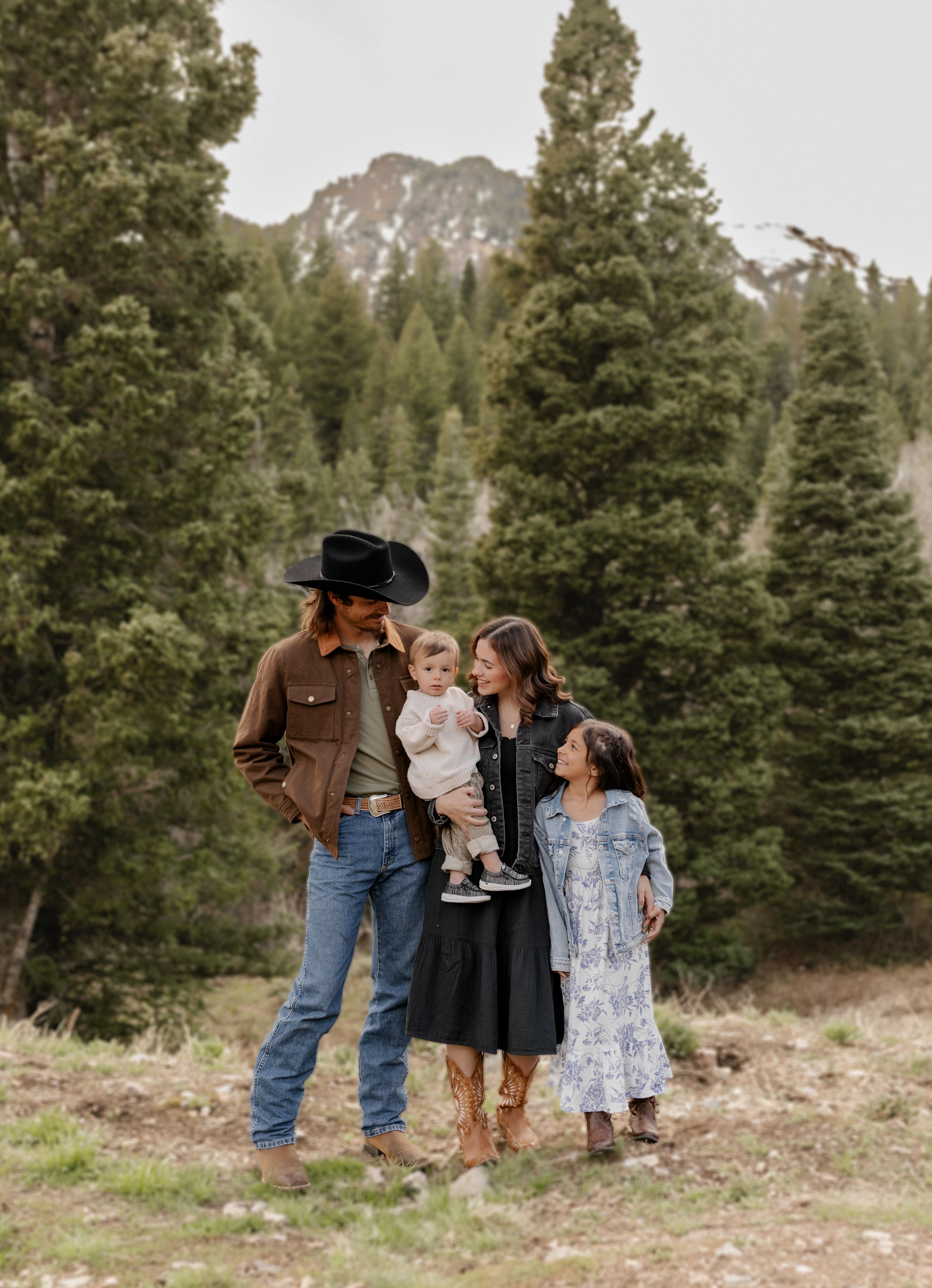A family in western attire poses together against a mountain backdrop with evergreen trees.