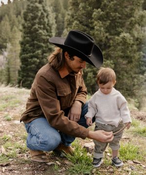 Someone in western wear kneels beside a small child in the mountains showing something interesting.