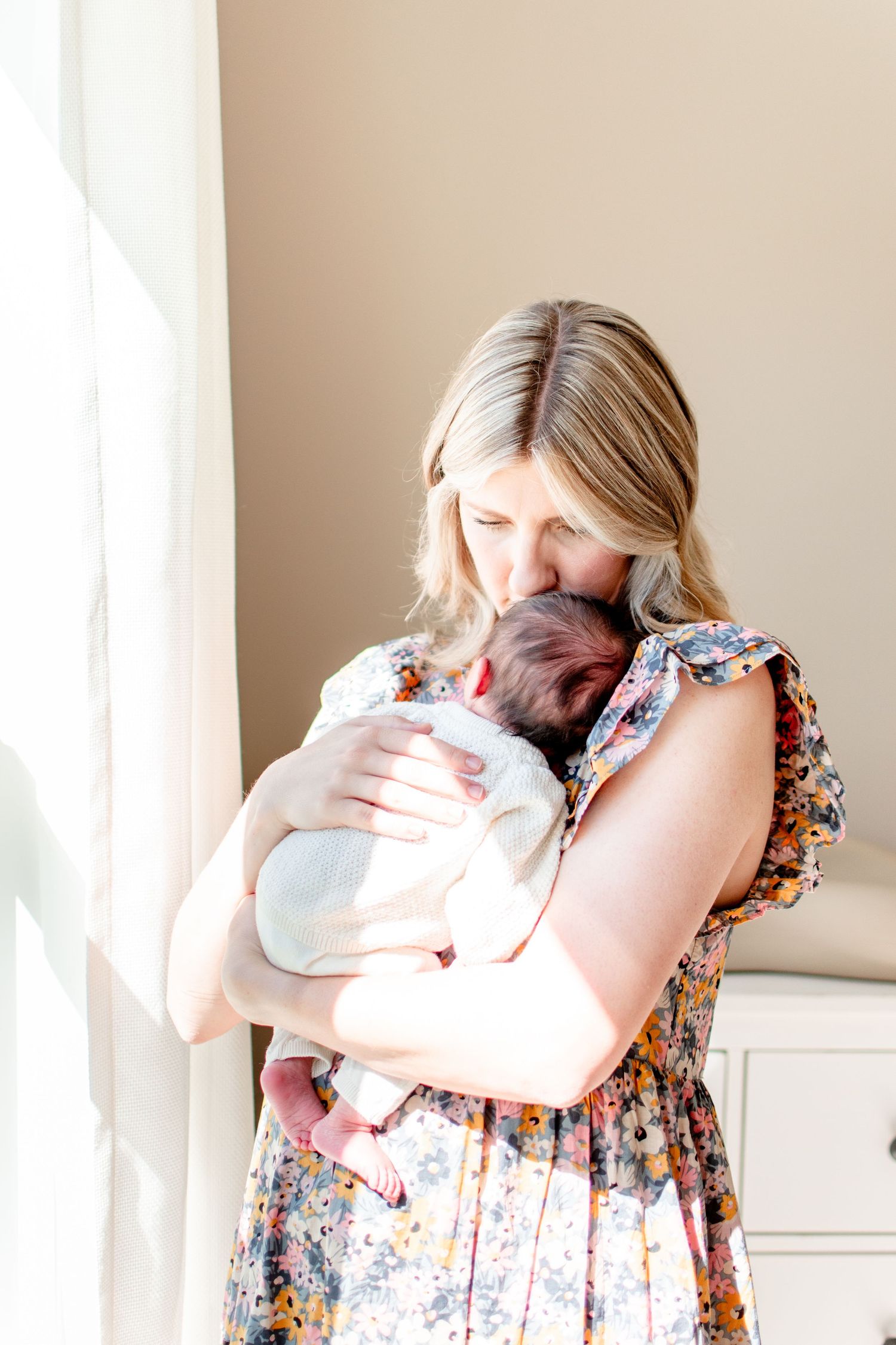 Mom kisses baby girl's forehead as they stand in the sunlight passing through the window of their home.