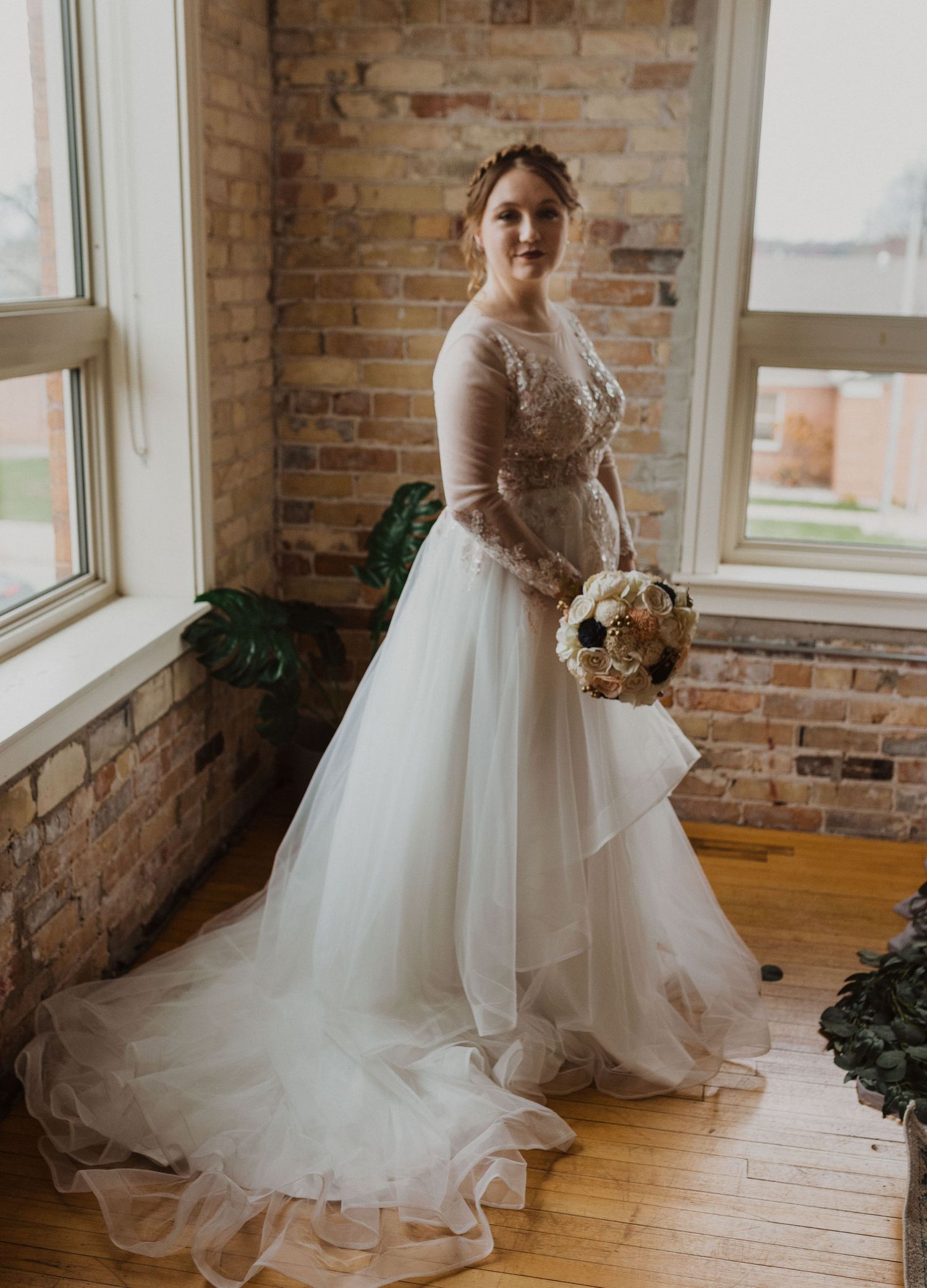 A bride holds her bouquet while showing off her flowing white wedding gown in a sunlit room.