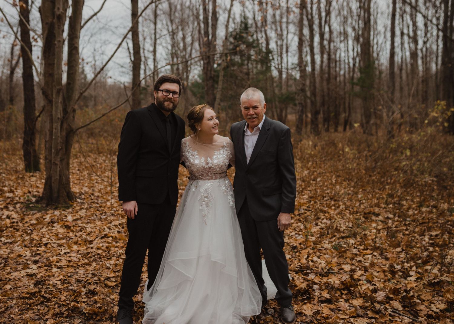 Wedding couple poses together in a wooded area with golden fallen leaves covering the ground.