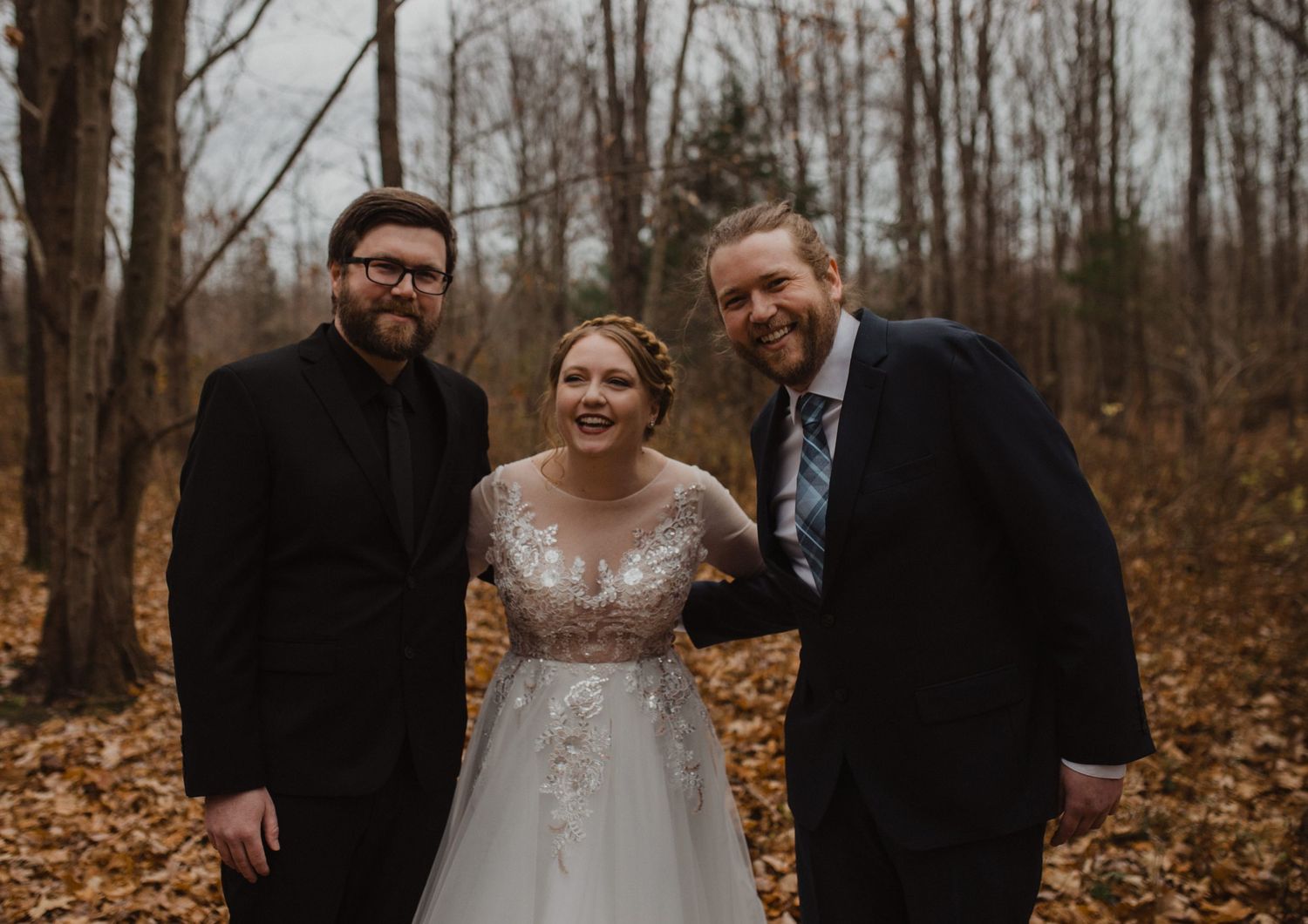 Wedding party stands together in a wooded setting with autumn foliage on the ground.
