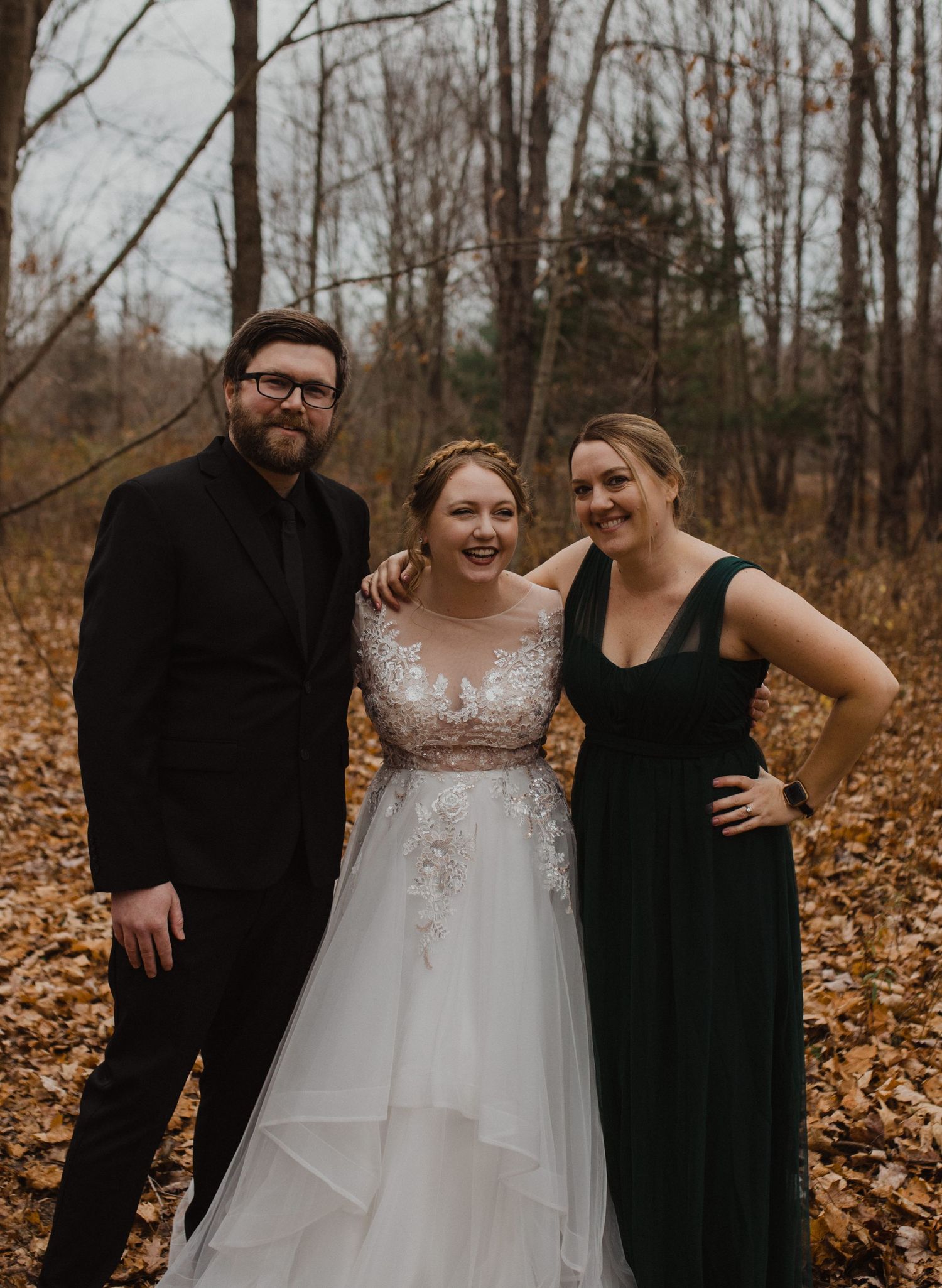 Wedding party members in black dresses gather around the couple in a fall forest setting.