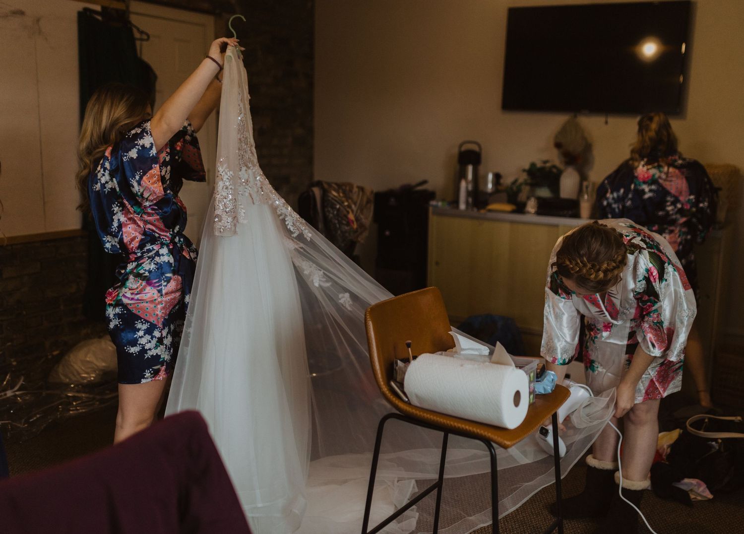 Bridesmaids in floral robes preparing white wedding dress.