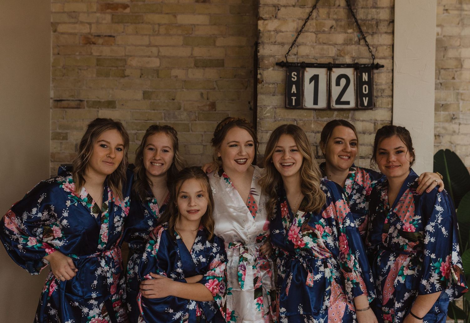 Wedding party in matching floral robes posing against brick wall.
